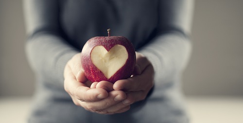 A person holding a red apple in their hands.There is a heart carved out of the apple.